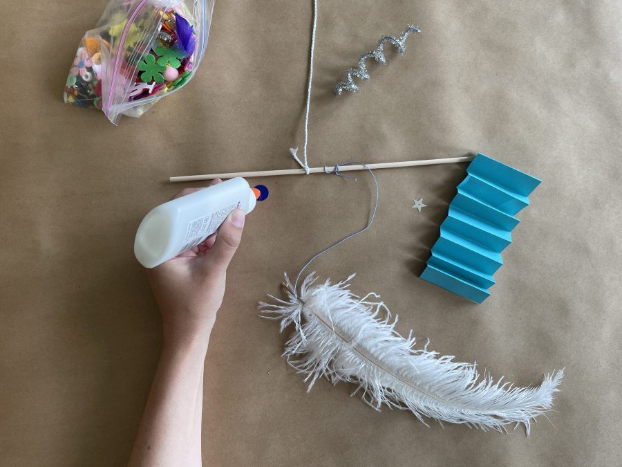 Single arm with hand holding glue, a wooden dowel, a white feather, and a clear plastic bag full of colorful knick knacks.