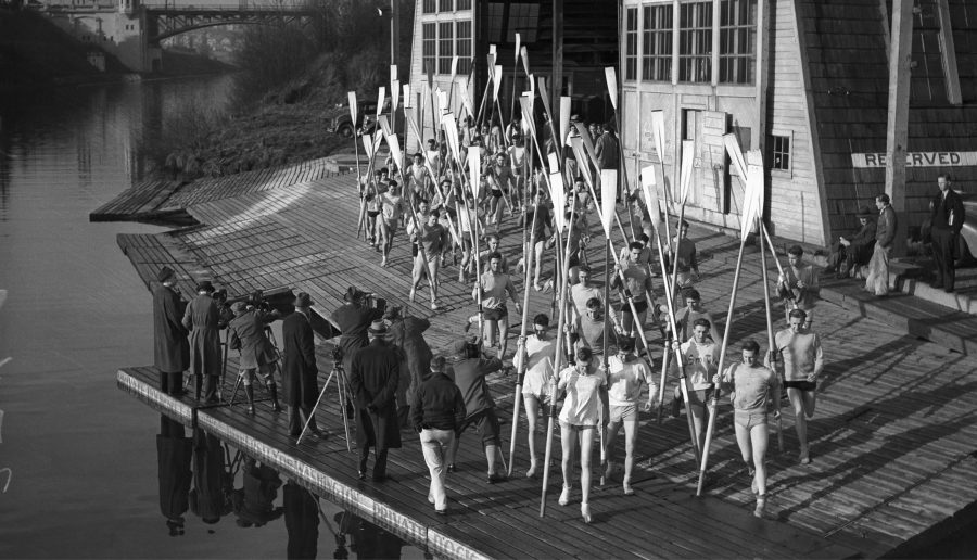 A historic black and white photo of the University of Washington Men's Crew Team carrying oars from the boathouse to the water.